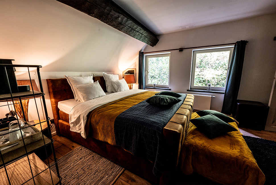 Le lit d'une chambre du Domaine de Béronsart avec des couvertures ocre et bleu foncé et une fenêtre offrant une vue sur le jardin