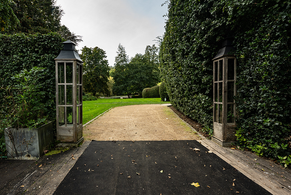 Entrée du Domaine de Béronsart avec deux grandes lampes brunes, un tapis noir au sol et des haies de part et d'autre de l'allée centrale