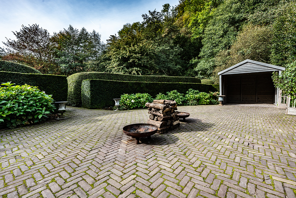 L'espace extérieur de la Serre du Domaine de Béronsart avec des bois de chauffage, un abri de jardin, des haies et des plantes vertes